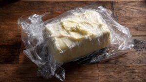 A block of soft, pale yellow butter partially wrapped in clear plastic on a rustic wooden surface